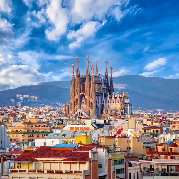 Stadtpanorama von Barcelona mit der emporragenden Sagrada Familia vor einem Gebirgszug im Hintergrund.