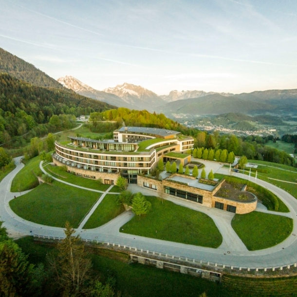 Luftaufnahme eines hufeisenförmigen Hotelkomplexes mit Rasenflächen inmitten einer hügeligen Waldlandschaft.