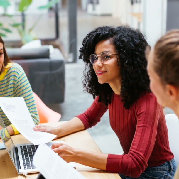 Drei junge Frauen sitzen mit Arbeitsunterlagen zusammen an einem Holztisch in einem Co-Working-Space.