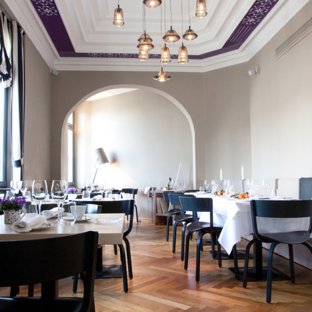 Heller Speisesaal eines edlen Restaurants mit schwarzen Holzstühlen an eingedeckten Tischen in einem klassischen Altbau mit hohen Decken und Holzparkett.