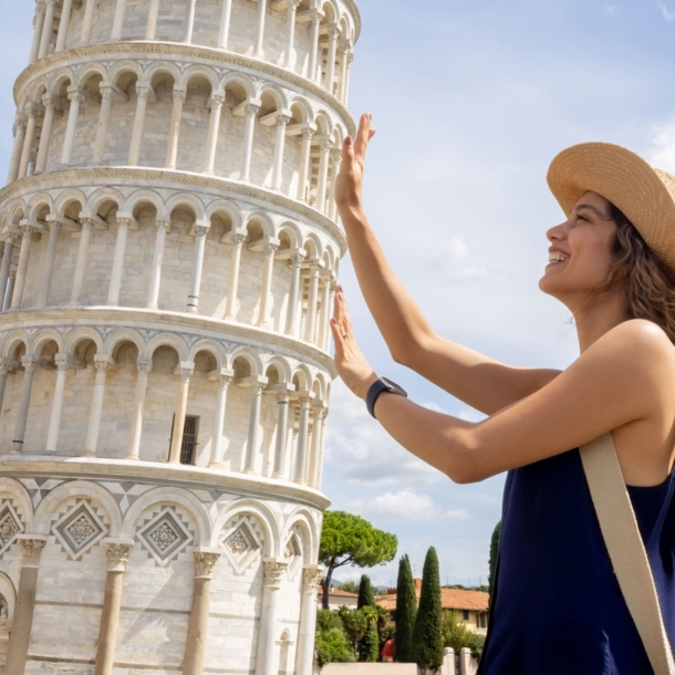 Profilansicht einer jungen Frau mit Strohhut, die aus der fotografischen Perspektive den Schiefen Turm von Pisa mit ihren Händen zu halten scheint.