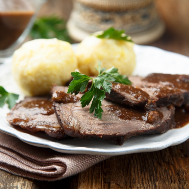 Sauerbraten mit brauner Soße an Klößen, angerichtet auf einem weißen Teller auf einem Holztisch.