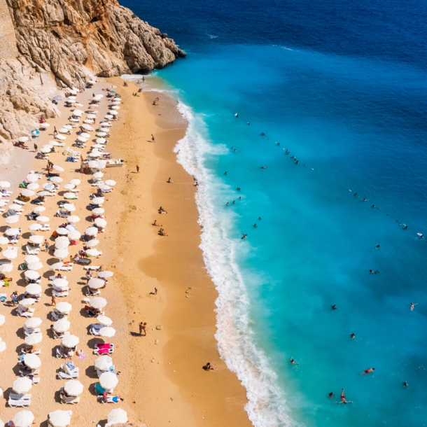 Luftaufnahme eines goldenen Sandstrandes mit Sonnenschirmen in einer Felsbucht mit Personen im türkisblauen Wasser.