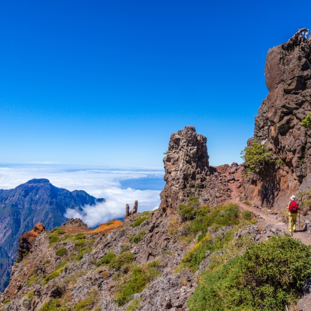 Eine Person auf einem Wanderweg auf einem Bergkamm mit rötlichen Felsformationen oberhalb der Wolkendecke unter blauem Himmel.