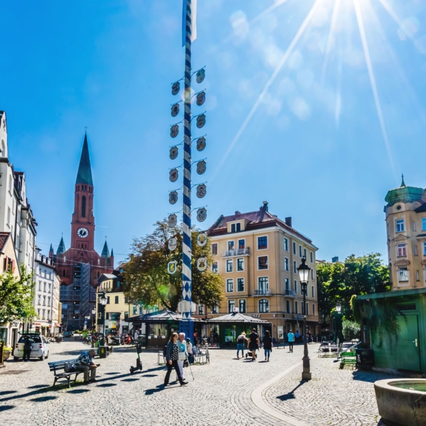 Ein Stadtteilplatz mit Kopfsteinpflaster und einem Maibaum in der Mitte bei strahlendem Sonnenschein.