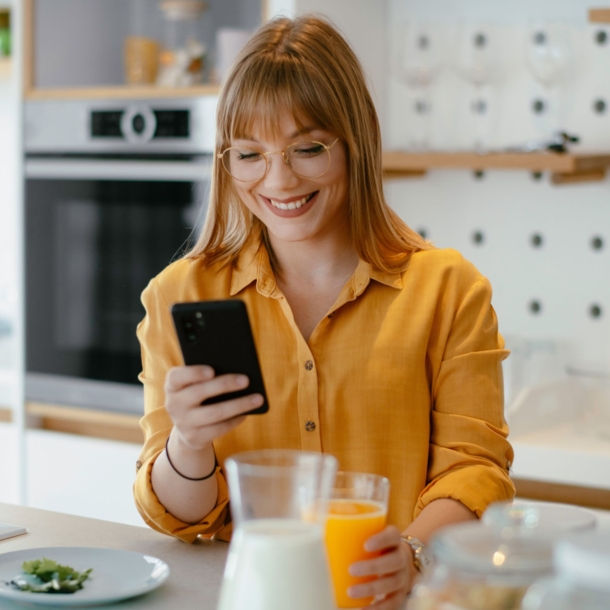 Eine junge Frau in gelbem Hemd steht mit einem Glas Orangensaft in der Hand in der Küche und schaut lächelnd auf ihr Smartphone.