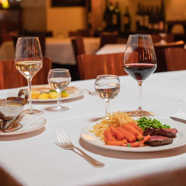 Zwei auf weißen Tellern angerichtete Speisen stehen neben Weingläsern auf einem Tisch mit weißer Stofftischdecke in einer Brasserie.