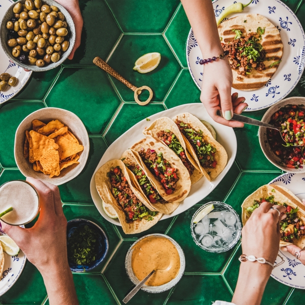 Aufnahme eines Tisches von oben, an dem mehrere Personen sitzen. Auf dem Tisch stehen verschiedene Teller mit mexikanischem Essen wie Tacos, Dips, Nachos und mehr.