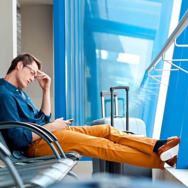 Ein Mann, der am Flughafen auf einer Bankreihe mit ausgestreckten Beinen sitzt. In einer Hand hält er ein Smartphone, die andere Hand ist an seiner Schläfe. Neben ihm steht ein Koffer.