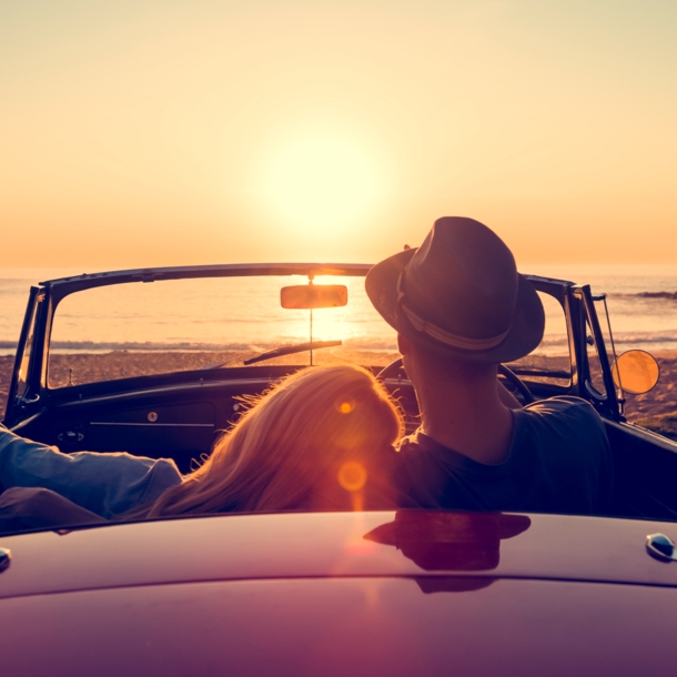 Rückansicht eines Paares in einem Cabriolet an einem Sandstrand bei Sonnenuntergang, sie hat den Kopf auf seine Schulter gelehnt.