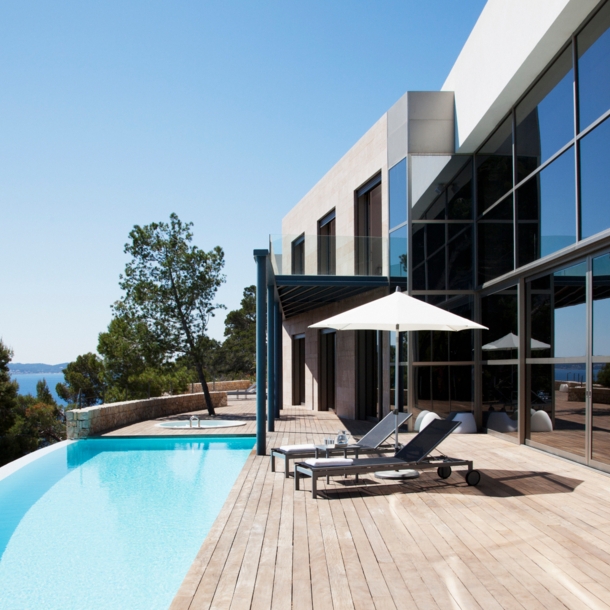 Ein Haus mit großer Glasfensterfront und angeschlossener Holzterrasse mit Pool. Auf der Terrasse befinden sich ein Sonnenschirm und zwei Liegen.