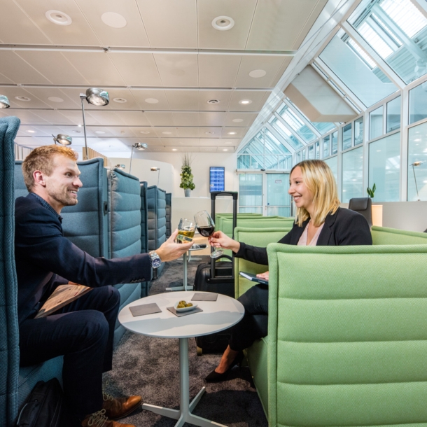 Ein Mann und eine Frau sitzen sich gegenüber auf Sesseln in einer Flughafenlounge und stoßen mit Weingläsern an.