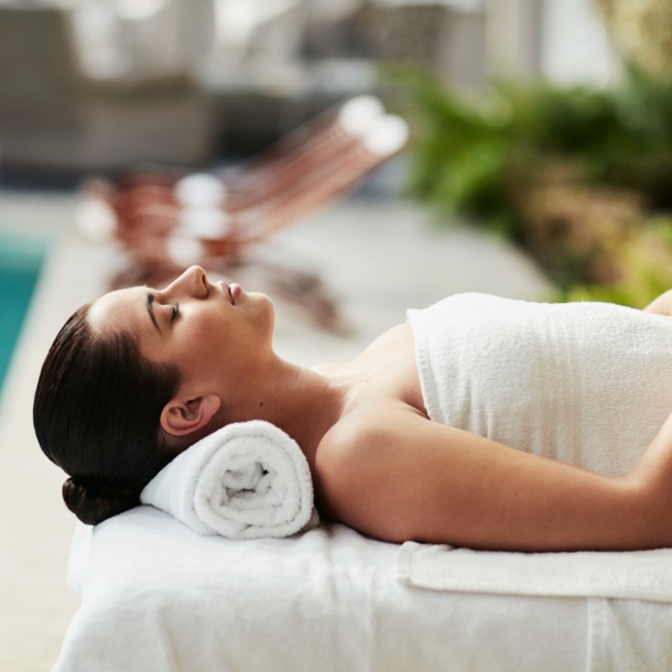 Eine Frau entspannt in einem weißen Handtuch auf einer Massageliege an einem Hotelpool.