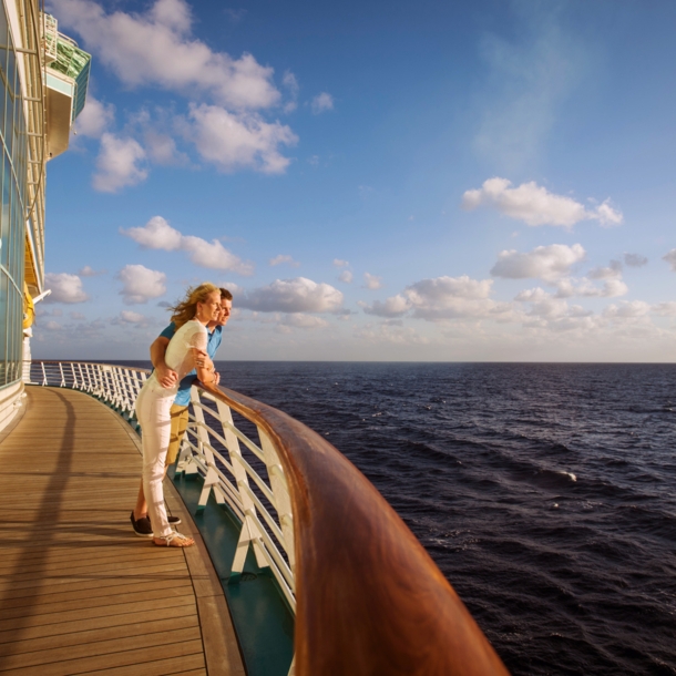 Ein Paar steht an der Reling eines Kreuzfahrtschiffes und schaut aufs Meer im Sonnenschein.