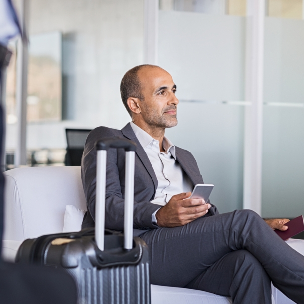 Ein Geschäftsmann sitzt in einem Sessel in einer Flughafenlounge, neben ihm steht ein Koffer.