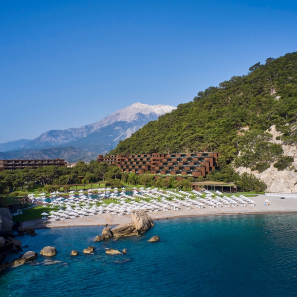 Ein Hotelresort in einer mediterranen Bucht mit Sandstrand, im Hintergrund eine Berglandschaft.