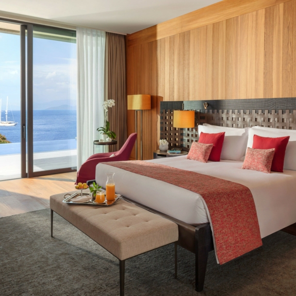 Modern eingerichtete Suite des Hotels Mandarin Oriental Bodrum mit einem großen Bett, einer Sitzbank, Arbeitstisch und raumhohen Fenstern mit Meeresblick.