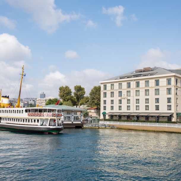 Gebäude mit neoklassizistischer Fassade am Ufer eines Gewässers mit Schiff im Vordergrund, im Hintergrund die Skyline von Istanbul.