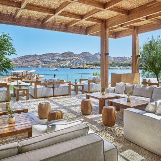 Loungebereich mit grauen Sesseln und Holztischen auf einer überdachten Terrasse an einer mediterranen Bucht.