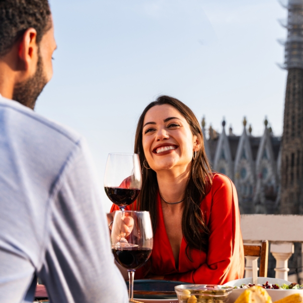 Eine Frau sitzt gegenüber von einem Mann an einem Tisch und hält lächelnd ein Glas Rotwein in der Hnad. Im Hintergrund die Sagrada Familia von Barcelona.