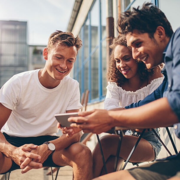 Eine Gruppe von Menschen sitzt zusammen auf dem Balkon und schaut gemeinsam etwas auf dem Smartphone.