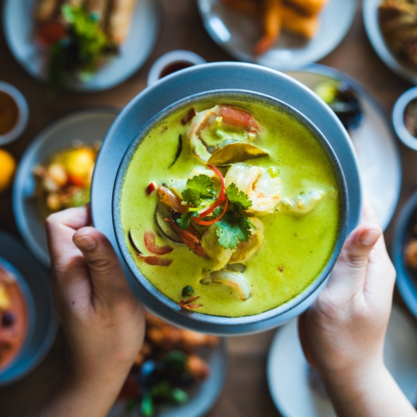 Zwei Hände halten eine Schale mit einem grünen Curry-Gericht über einen gedeckten Tisch mit vielen weiteren Speisen.