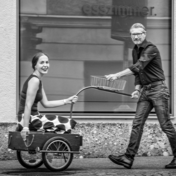 Ein Mann zieht eine lächelnde Frau in einem Fahrradanhänger auf einem Gehweg vor einer Fensterfront.