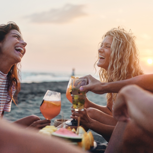 Eine Gruppe von vier fröhlichen Personen sitzt am Meer bei Sonnenuntergang mit Drinks und Snacks.