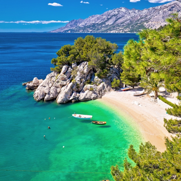 Kleine Sandbucht mit Fischerbooten im türkisblauen Meer, umgeben von Felsen und Kiefernwald.