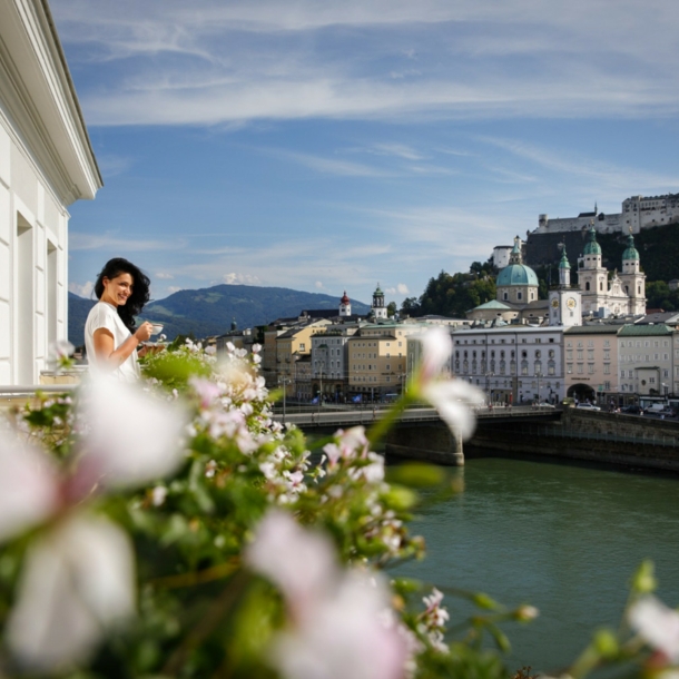 Eine Frau steht mit Kaffeetasse auf einem Balkon am Fluss, ihr gegenüber die Salzburger Altstadt mit Burgfestung.