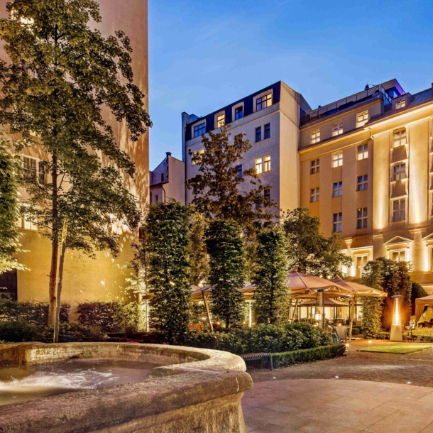 Imposantes, beleuchtetes Hotelgebäude mit barocker Fassade, im Vordergrund ein Springbrunnen mit Löwenkopf.