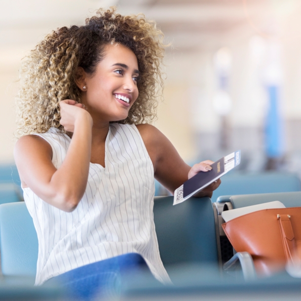 Eine junge, lächelnde Frau sitz mit Reisepass in der Hand in einer Flughafenlounge.