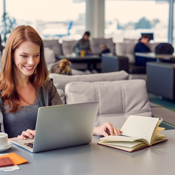 Eine lächelnde Frau arbeitet an ihrem Laptop an einem Tisch in einer Flughafen-Lounge.