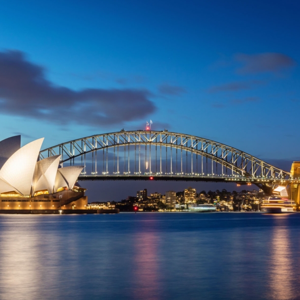 Sydney Harbour Bogenbrücke hinter dem Sydney Opernhaus am Wasser vor der erleuchteten Skyline Sydneys bei Nacht.