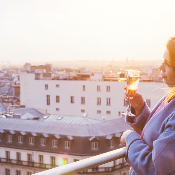 Eine Frau, die bei Sonnenuntergang in Paris auf einem Balkon steht und ein Glas Sekt in der Hand hält.