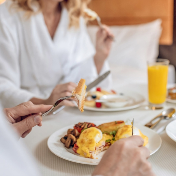 Nahaufnahme von zwei gesichtslosen Personen in weißen Bademänteln, die an einem eingedeckten Tisch in einem Hotelzimmer frühstücken.