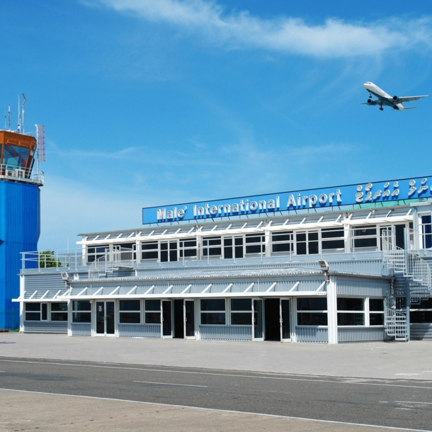 Ein Flugzeug fliegt am blauen Himmel über einen kleines Flughafengebäude mit blauem Kontrollturm.