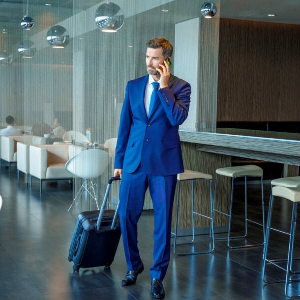 Ein Geschäftsmann im blauen Anzug und mit Trolley telefoniert stehend in einer modernen Flughafen-Lounge.