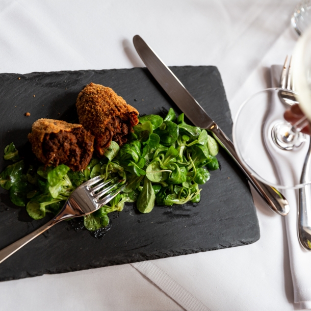 Aufsicht eines panierten Fleischgerichtes an Feldsalat auf einer Schieferplatte auf einem edel gedeckten Tisch, im Vordergrund eine Hand mit einem Glas Weißwein.