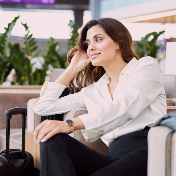 Eine Frau in Business-Kleidung sitzt in einem hellen Lounge-Sessel. Neben ihr steht ein Koffer, auf dem Tisch vor ihr befinden sich Reisedokumente und eine Tasse mit Untersetzer.