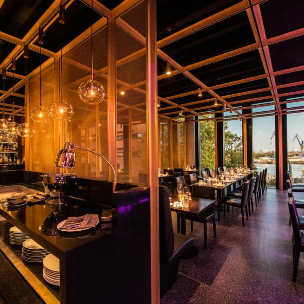 Offener Speiseraum eines stylischen Restaurants mit Blick auf Hafenareal durch Panoramafenster.