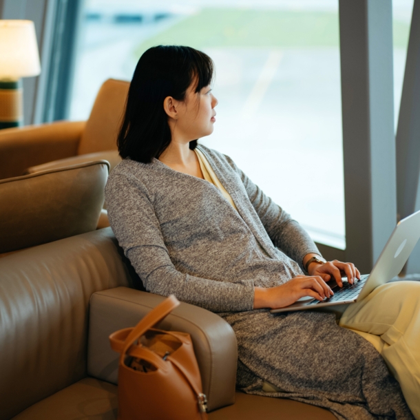 Eine Frau sitzt mit Laptop in einem Ledersessel am Fenster einer Flughafen-Lounge und schaut aufs Rollfeld.