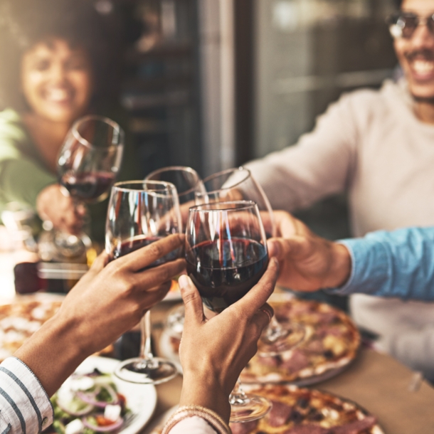 Eine Gruppe von lächelnden Menschen, die an einem Tisch sitzt, auf dem Teller mit Pizza und anderem Essen stehen, und die mit jeweils einem Rotweinglas anstoßen.