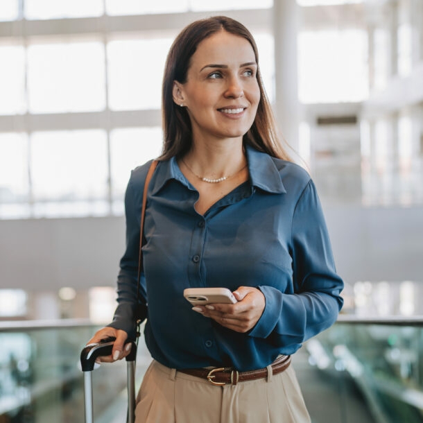 Eine lächelnde, elegant gekleidete Frau mit Smartphone und Trolley auf einer Rolltreppe in einem Flughafenterminal.