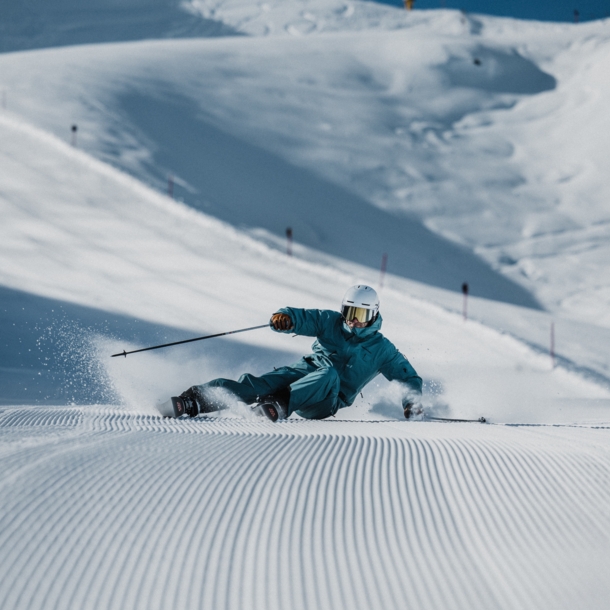 Ein Skifahrer mit Helm im türkisen Schneeanzug bei der Abfahrt auf einer Piste.