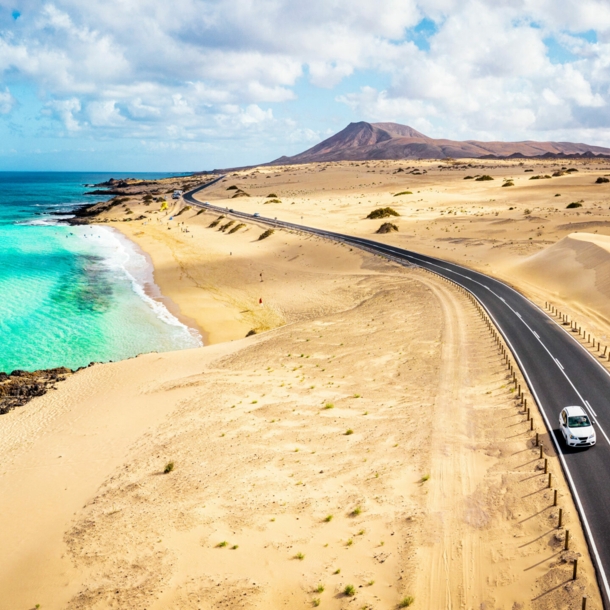 Ein weißes Auto fährt auf einer Schnellstraße durch eine Wüstenlandschaft am türkisblauen Meer.