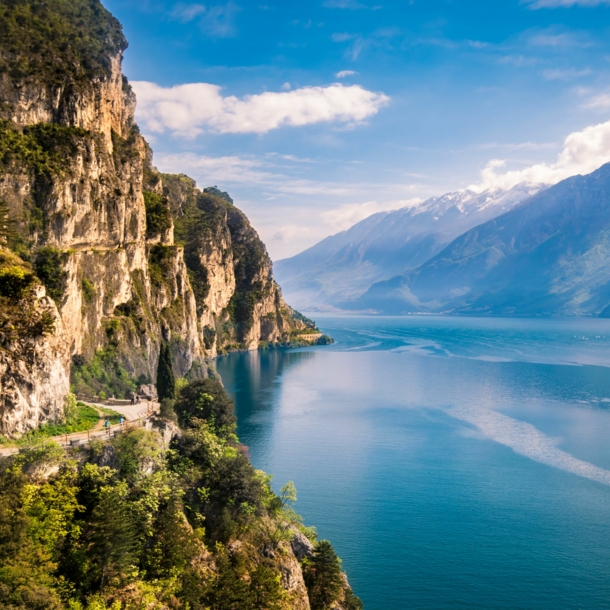 Bewachsene Felswand nahe Riva del Garda direkt am Gardasee mit Gipfeln auf der anderen Seeseite im Hintergrund.