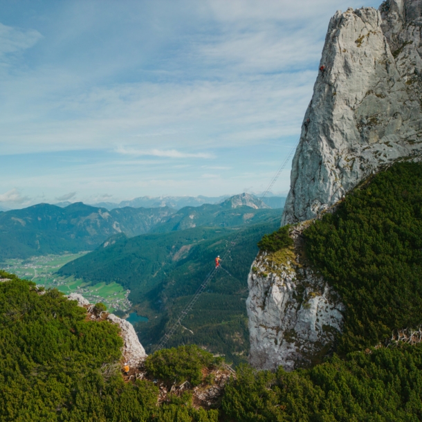 Eine Person auf einer Leiter, die hoch über einem Abgrund zu einem hohen Felsen führt, im Hintergrund Berge und Himmel.