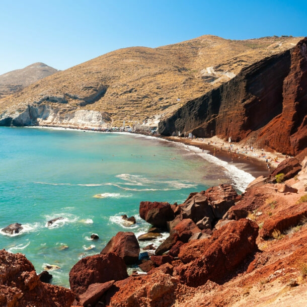 Meeresbucht mit Booten im türkisblauen Wasser, umgeben von roten Felsklippen an rotem, belebtem Sandstrand.