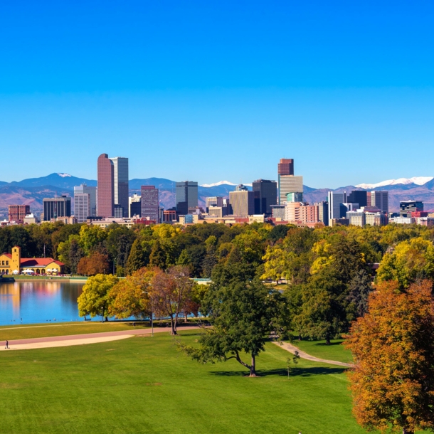 Skyline von Denver unter blauem Himmel vor Gebirgszug, im Vordergrund eine Parkanlage mit Teich und grünen Bäumen.